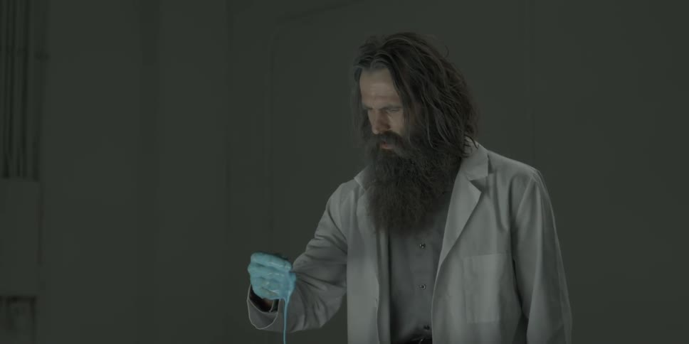 A man in a lab coat raises and looks at some blue liquid covering his hand
