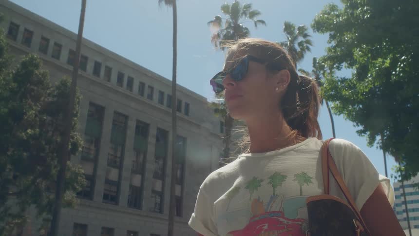 A woman wearing sunglasses stands on a street near a building and palm trees