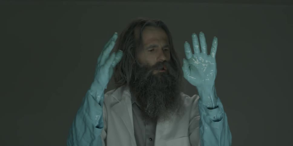 A disheveled man in a lab coat examines his hands that are covered in blue liquid