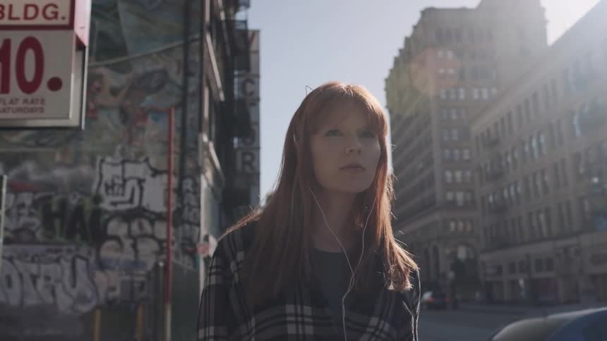A woman wears earbuds as she walks down a city sidewalk during the day