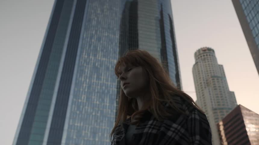 A red haired woman stands outside in front of tall buildings