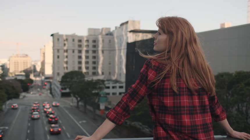 A woman leans into a railing on an overpass looking down on a city street