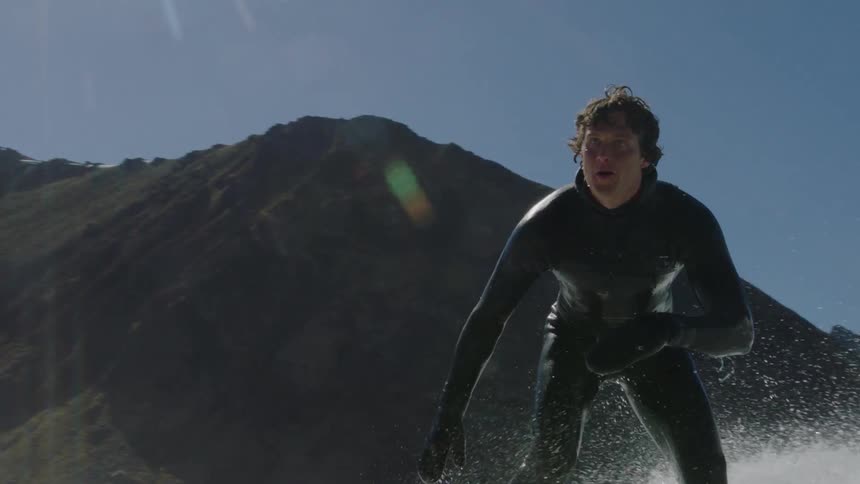 A surfer rides a wave along a rugged shore during the day
