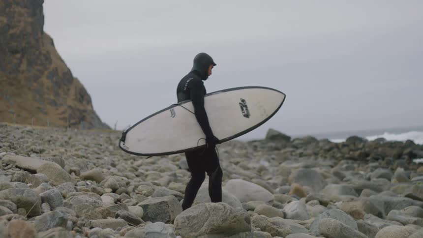 A surfer in a wetsuit carries his surfboard across rocks toward the sea