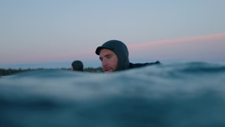 Two men in water suits swim on surfboards in water
