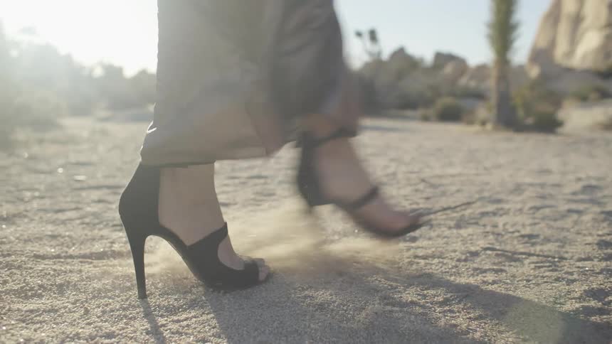 A woman in high heels and a dress is kicking up sand in the desert during the day