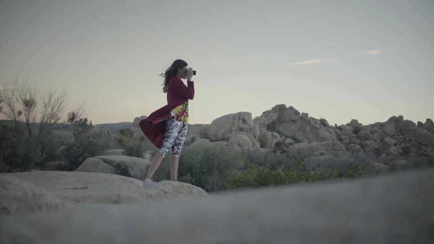 A woman is taking a photo as she stands on a boulder in the desert