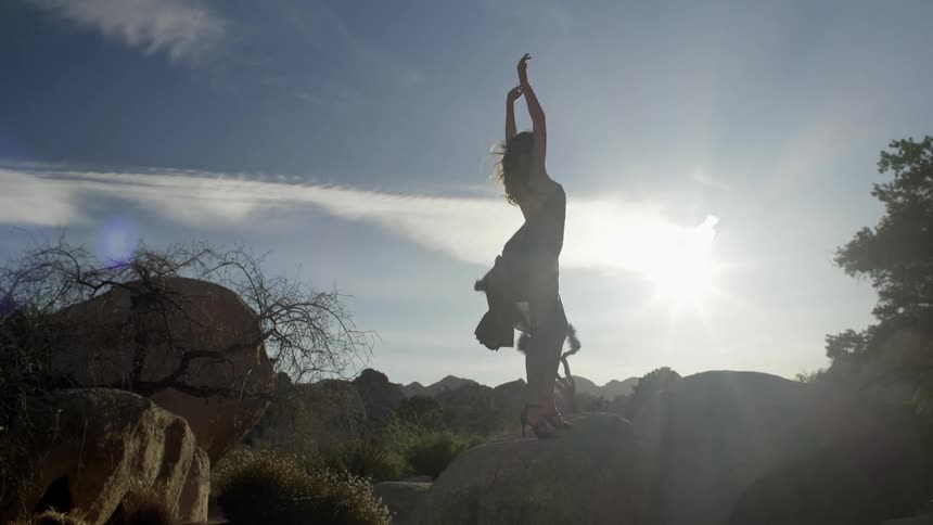 Woman stands on rocks as wind blows her hair and dress