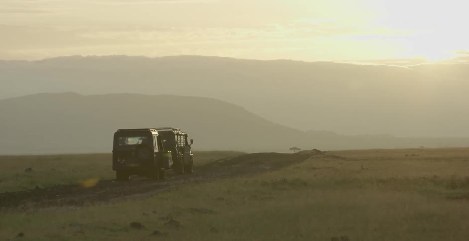 Two jeeps travel on a dirt path across an african savanna at sunset