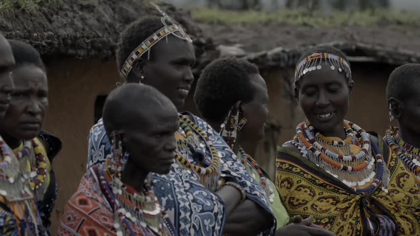 Maasai in costume stand outside a village talking and waiting