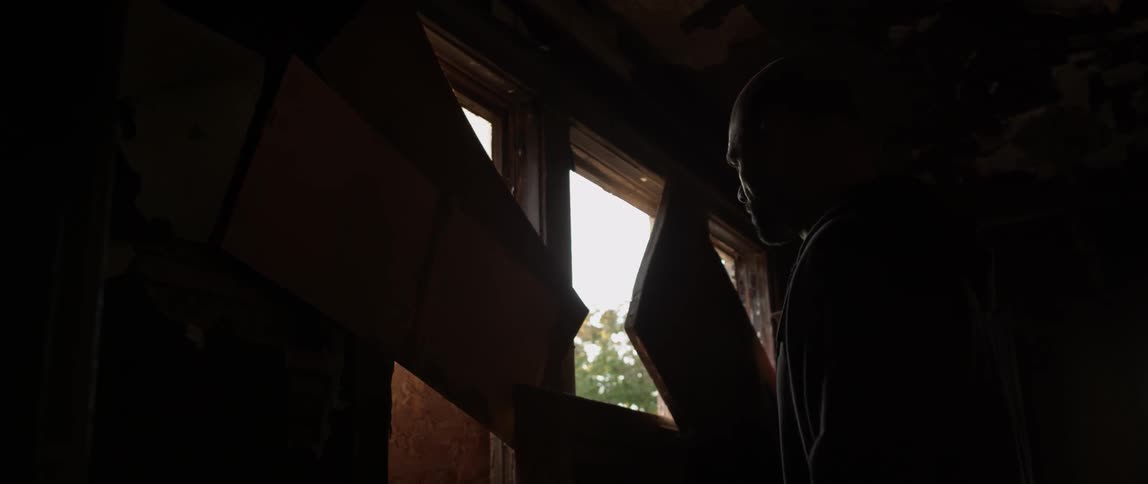 A man peers out through a boarded up window in an old building