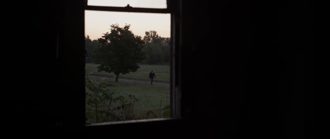 Outside the window of the dark house a person is walking down the trail
