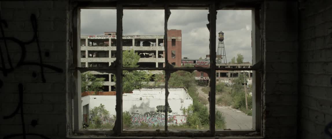 A broken window looks out on an abandoned parking garage and buildings during the day