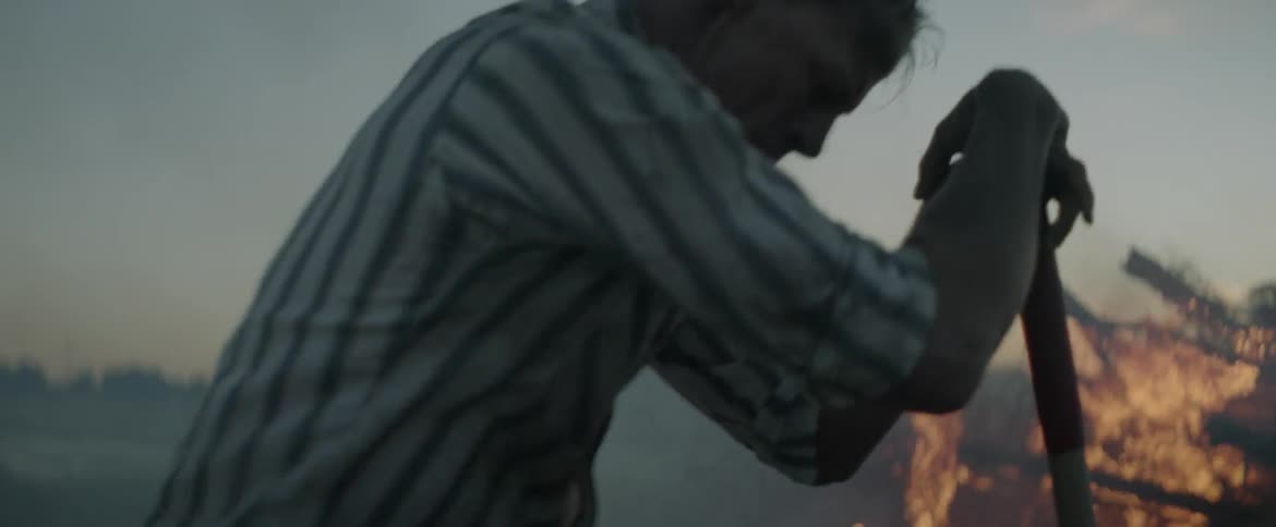 Man shoveling dirt onto a fire outdoors and then resting on his shovel looking tired