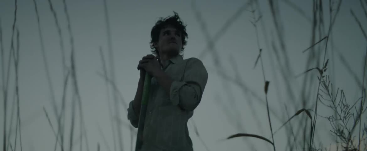A man stands resting his hands on a shovel on a windy evening