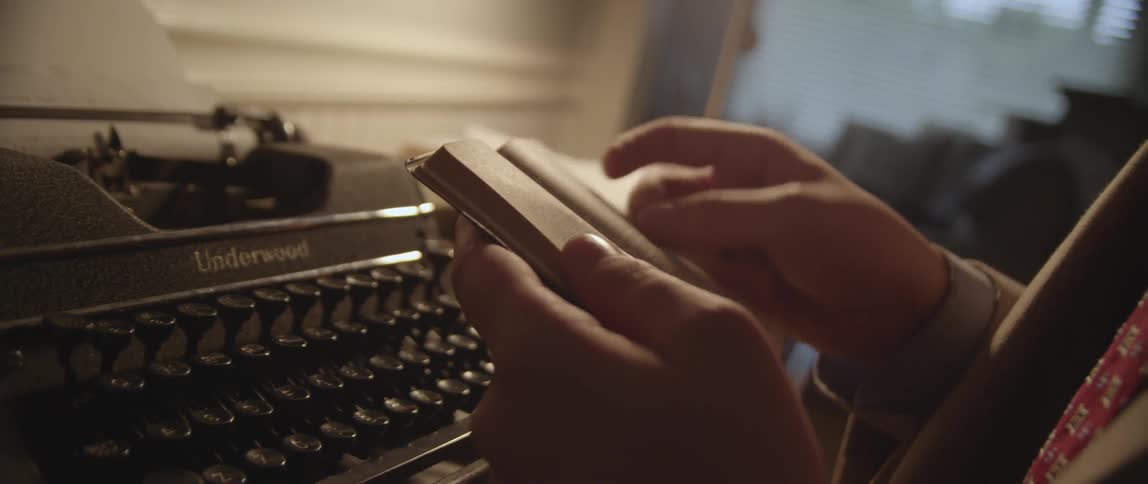 Someone sits in front of an old typewriter, turning the pages of a tiny book