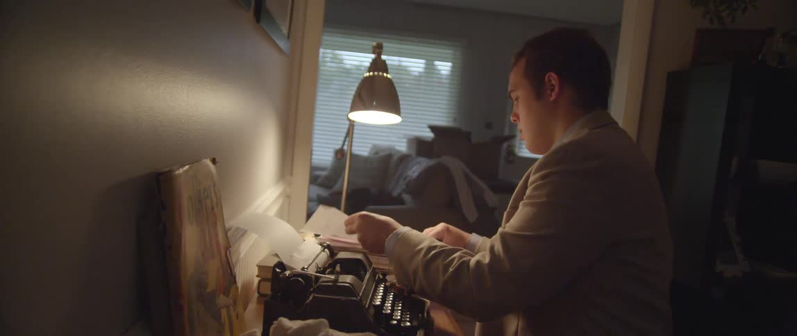A man sits at a typewriter and rummages through a stack of papers