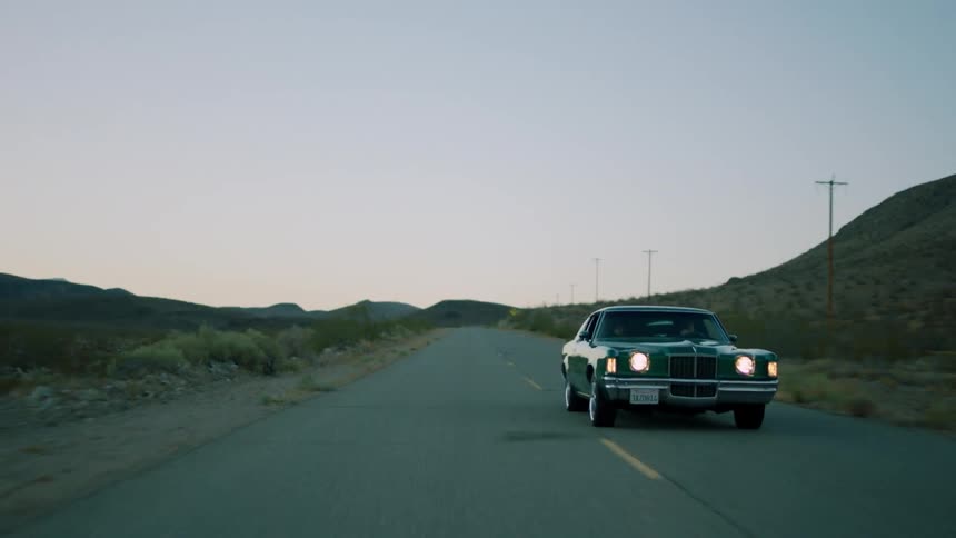 A late model american car is driving on a desert highway at dusk