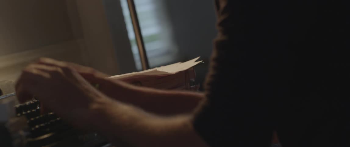 A man sits in front of an old typewriter, reviews some nearby papers then types
