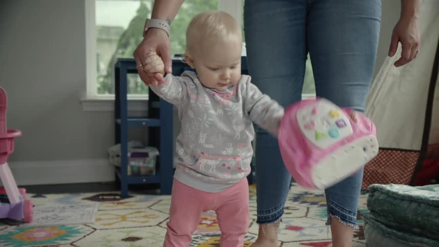 A toddler holds its mother's hand as it stands and plays in a living room