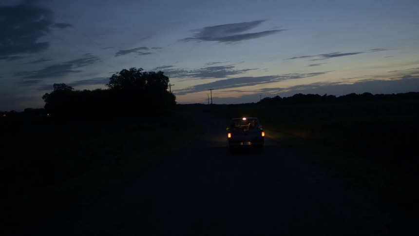 Three people in the back of a pickup truck drive down a dark road at sunset
