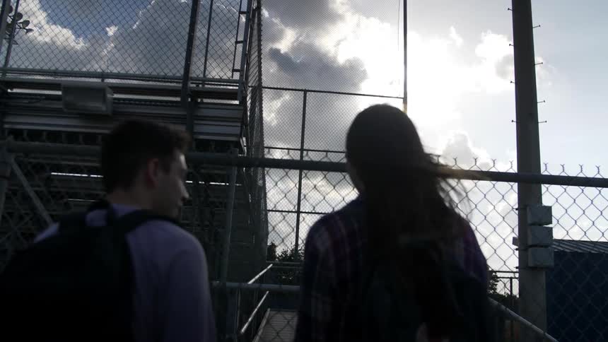 A young man and woman walk towards bleachers and run off