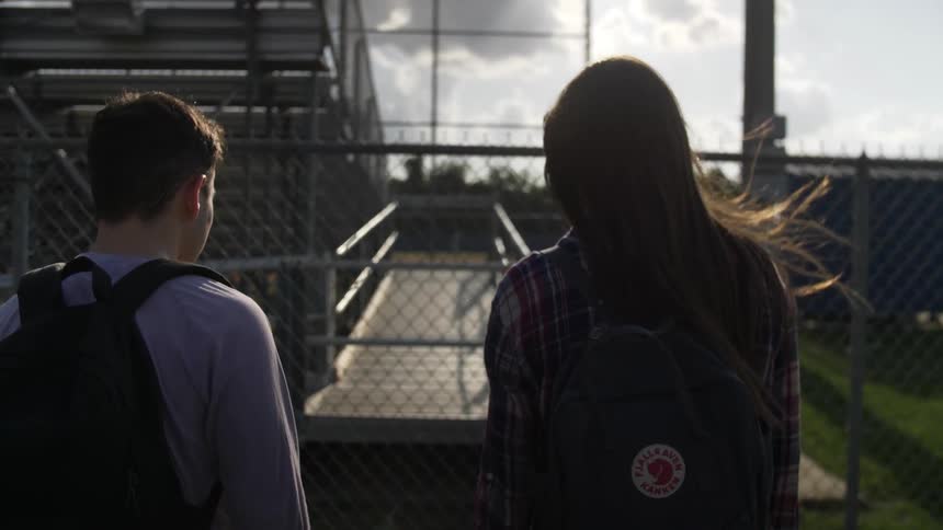 A teenage boy and girl are walking behind stadium stands at school during the day