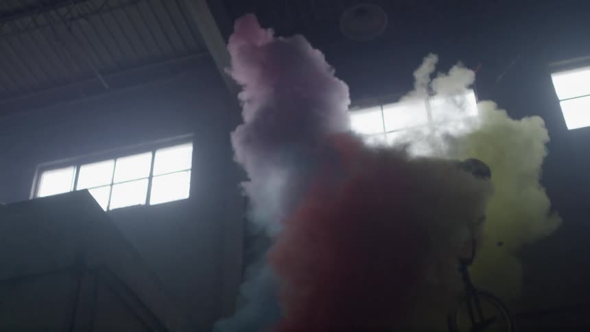 A male wearing a helmet while riding a bike in a warehouse proceeds to jump off a ledge while spinning around and goes through a cloud of blue yellow and red smoke as it explodes in the air