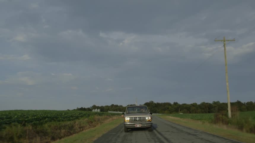 A truck is following behind another vehicle as they drive through a road with crops on either side