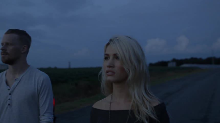 A man and woman stand outside a pickup truck staring down the road at dusk