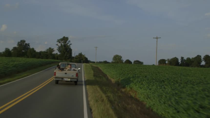 Two men rest in the back of a pickup truck driving on a road in farm country