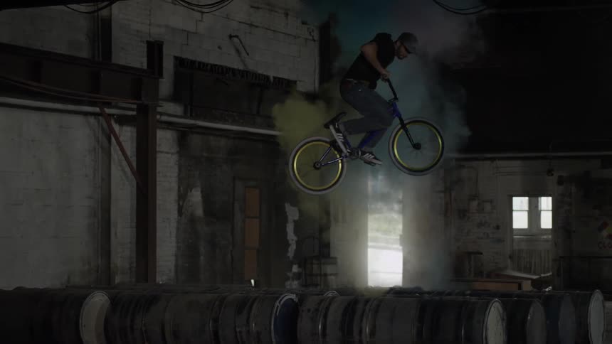 A man jumps a bicycle over rows of barrels as colored powder explodes