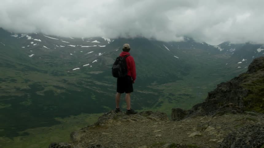 A man wearing a backpack stands on a ledge overlooking a valley and mountains