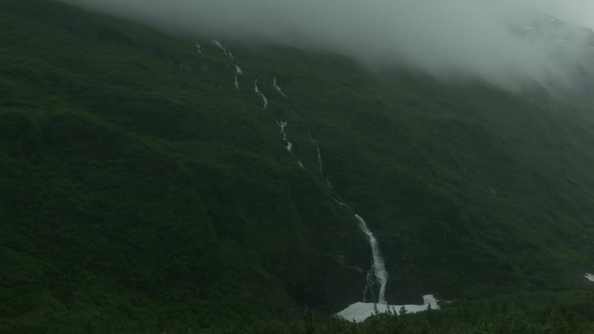 A waterfall cascades down a rolling hillside into a river on a misty day
