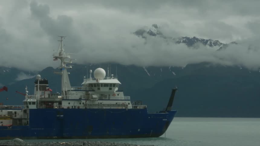 A commercial boat sits in the water in front of mountains covered by low clouds