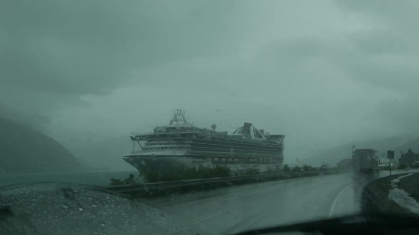 Vehicles traveling on a road pass by a cruise ship at anchor on a rainy day