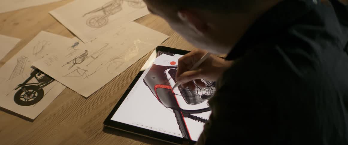 A man is working on sketches and a computer graphic of a motorcycle at a table