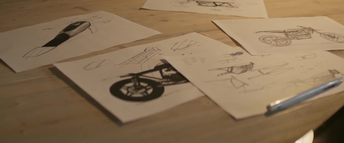 Sketches of a motorcycle are resting on a drafting table under lamplight