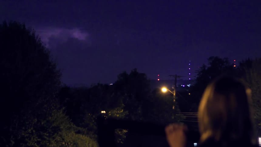 A woman is filming a lightning storm in the distance from a patio