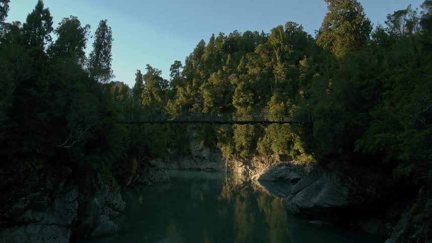 A drone flies under a bridge above calm green water between two forests