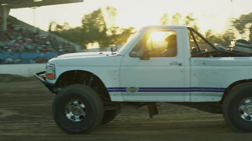 An audience watches pickup trucks race around a dirt track