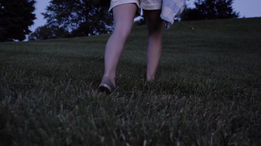 Woman walking through grassy field with fireflies at dusk
