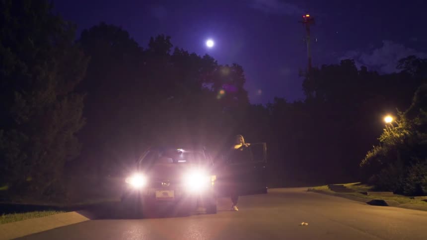A woman is out of her car and watching something off camera while leaning against the door