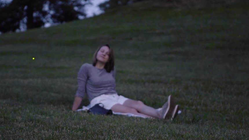Woman sits in grass with glowing fireflies nearby