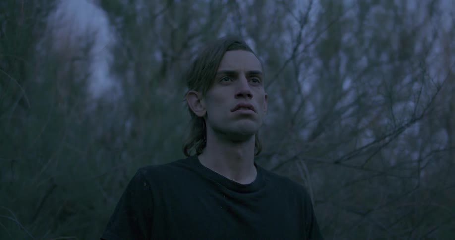 A man with long hair is standing in the woods looking at something