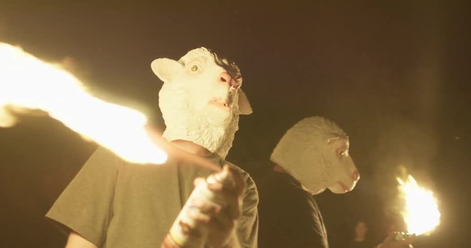 Two people in sheep masks are using aerosol cans to blow fire