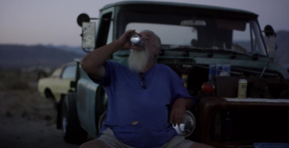 A man is sitting outside of his truck drinking a beverage from a can