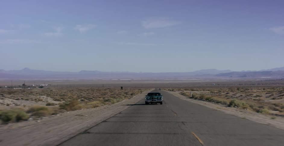 A late model american car is driving on a desert highway during the day