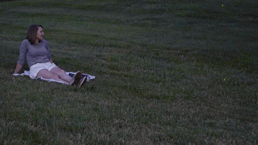 A lady is laying in the grass while fireflies are flying around her