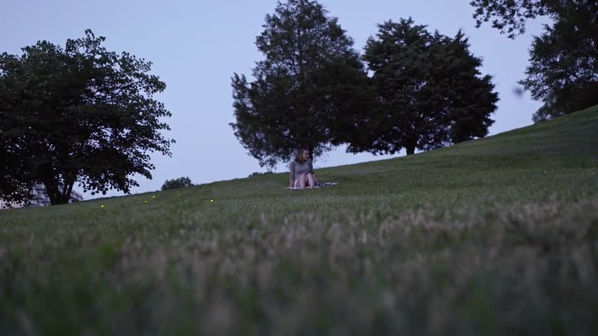 A woman sits in a park at dusk surrounded by glowing fireflies